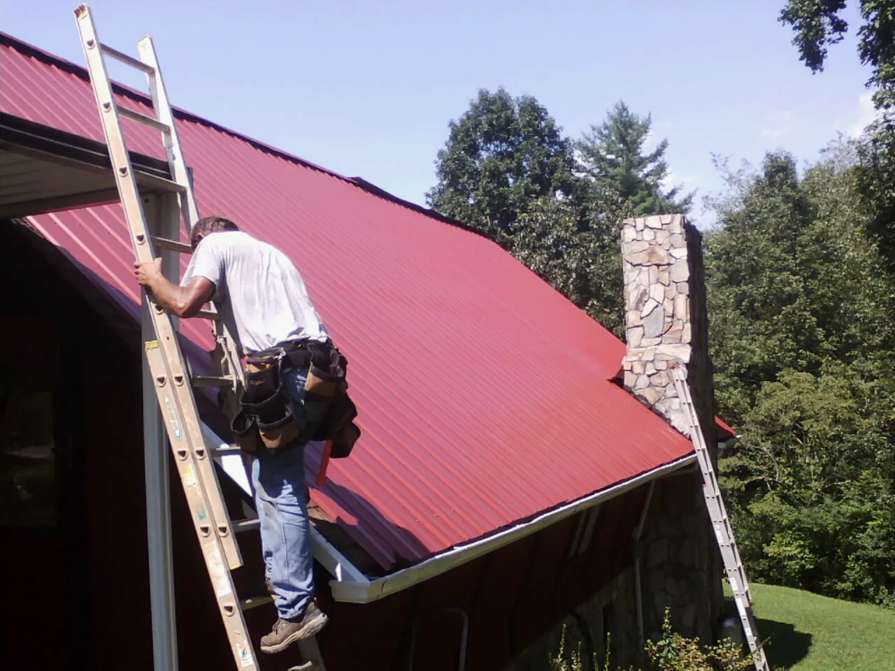Roofer on a ladder beside a red metal roof and stone chimney — Soffit & Fascia Repair by Roofing Brothers in Orrs Island